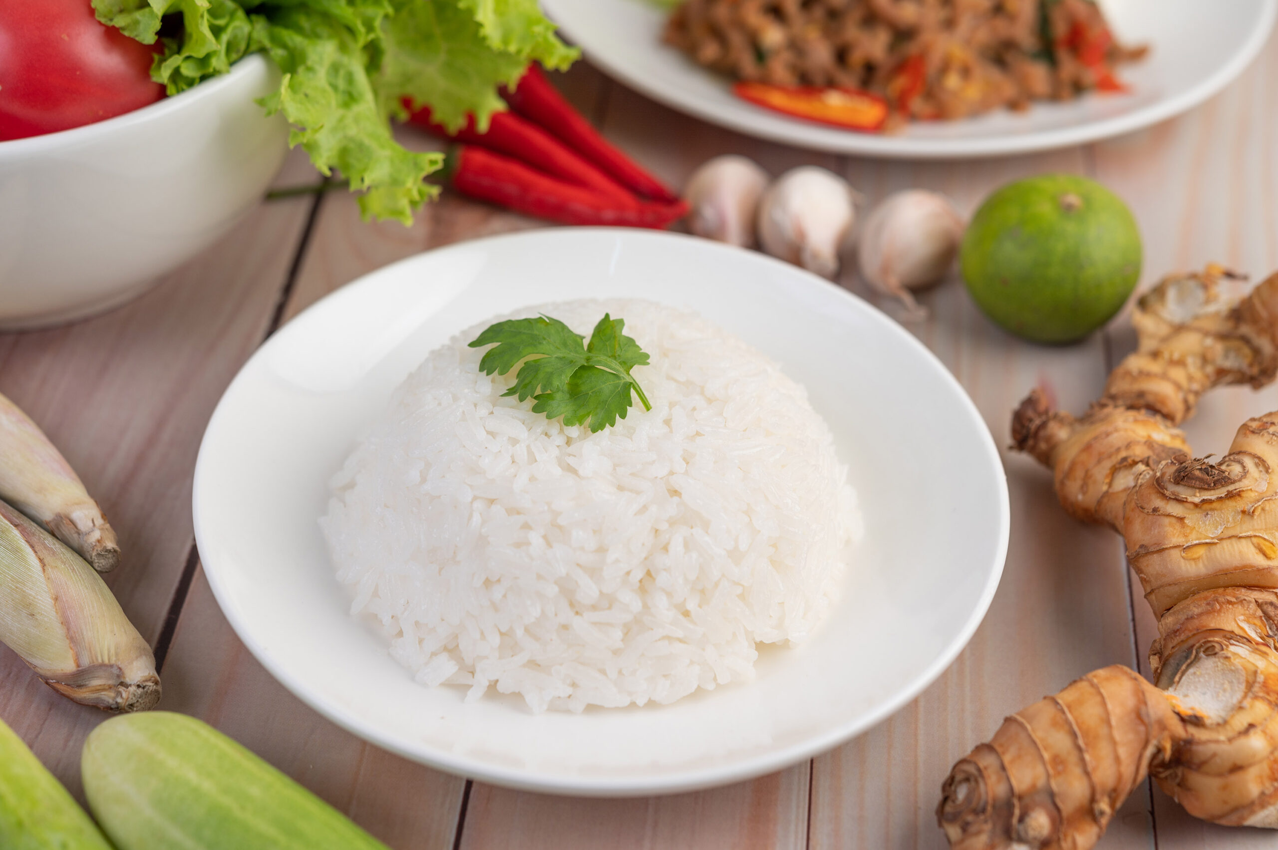 Rice cooked in a white dish with galangal, lemons, cucumbers, lemongrass, garlic, peppers, tomatoes and lettuce on a wooden floor.